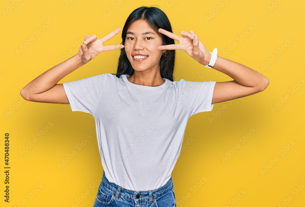 Beautiful young asian woman wearing casual white t shirt doing peace symbol with fingers over face, smiling cheerful showing victory Beautiful young asian woman wearing casual white t shirt doing peace symbol with fingers over face, smiling cheerful showing victory