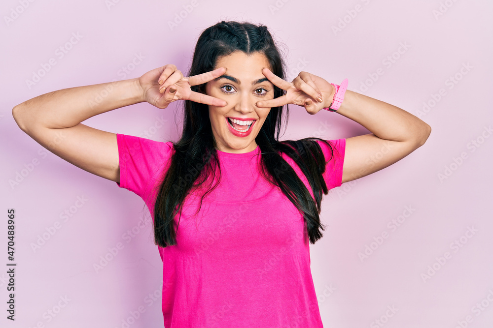 Young hispanic girl wearing casual pink t shirt doing peace symbol with fingers over face, smiling cheerful showing victory