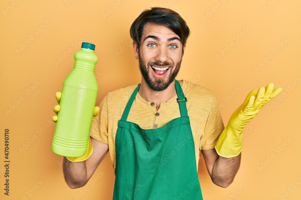 Young hispanic man wearing cleaner apron holding cleaning product celebrating victory with happy smile and winner expression with raised hands Young hispanic man wearing cleaner apron holding cleaning product celebrating victory with happy smile and winner expression with raised hands