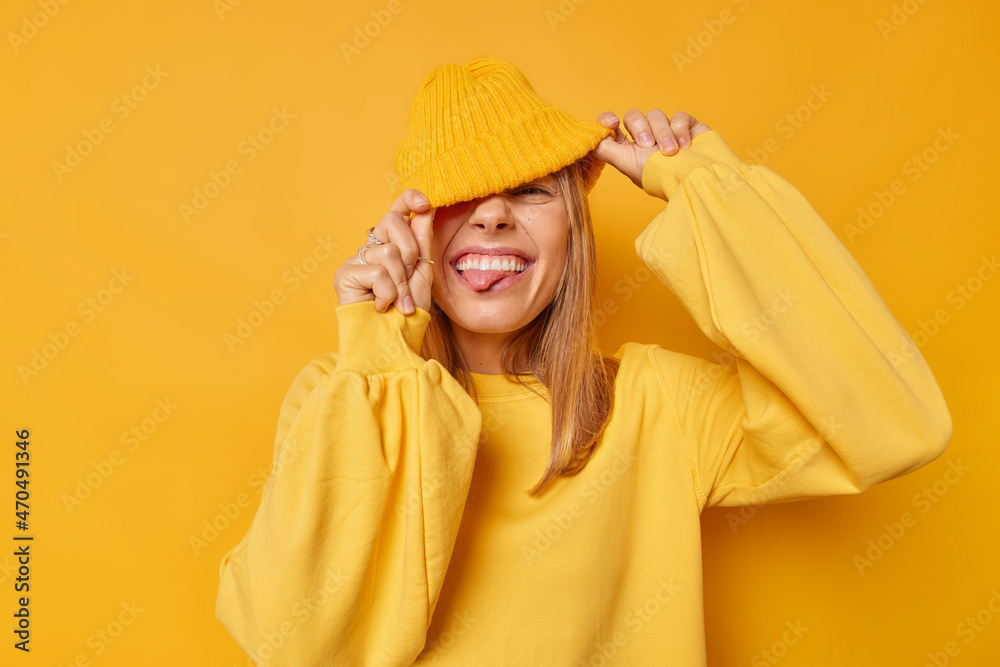 Joyful happy woman hides eyes with hat sticks out tongue has playful expression teases someone wears casual jumper isolated over yellow background squints happily shows true emotions. People and fun Joyful happy woman hides eyes with hat sticks out tongue has playful expression teases someone wears casual jumper isolated over yellow background squints happily shows true emotions. People and fun