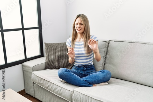 Asian young woman sitting on the sofa at home disgusted expression, displeased and fearful doing disgust face because aversion reaction. with hands raised