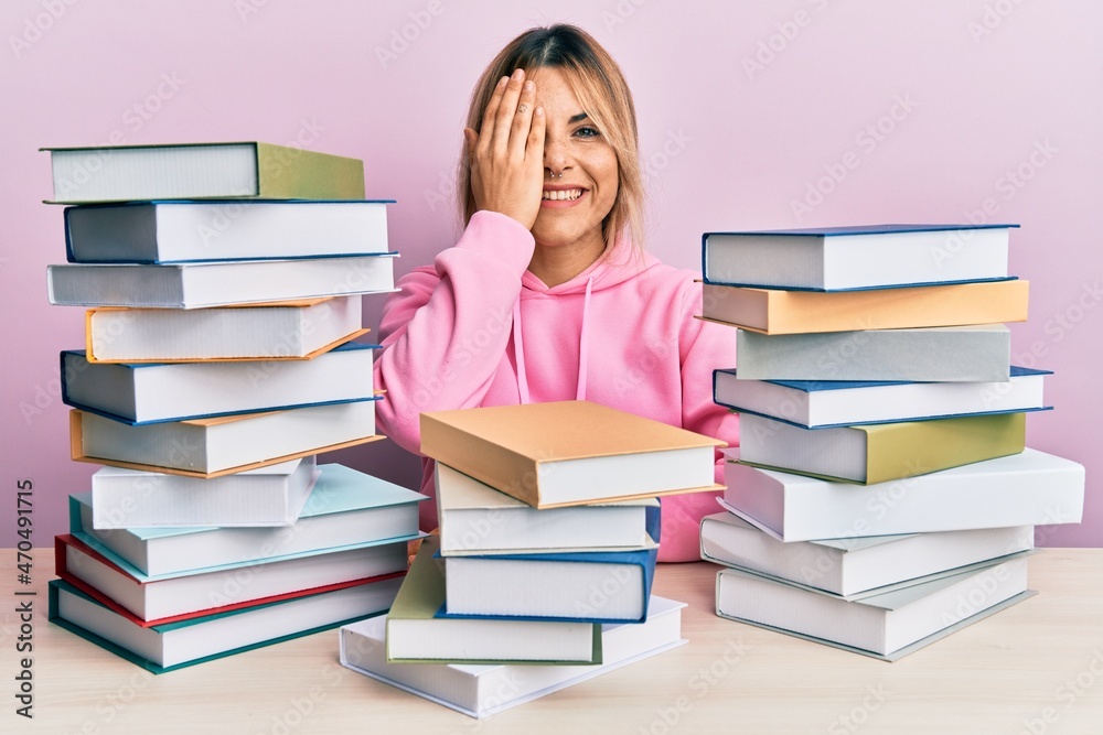 Young caucasian woman sitting on the table with books covering one eye with hand, confident smile on face and surprise emotion. Young caucasian woman sitting on the table with books covering one eye with hand, confident smile on face and surprise emotion.