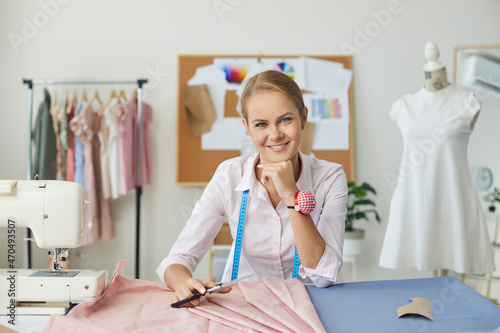Portrait of seamstress or tailor at work. Happy beautiful woman who works as dressmaker sitting at table with scissors and fabric in modern studio, resting chin on hand, looking at camera and smiling