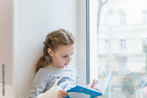 Cute little girl reading book on windowsill.Portrait of beautiful little European girl having concentrated look, resting on windowsill.studying indoor concept, clever schooler, daytime.