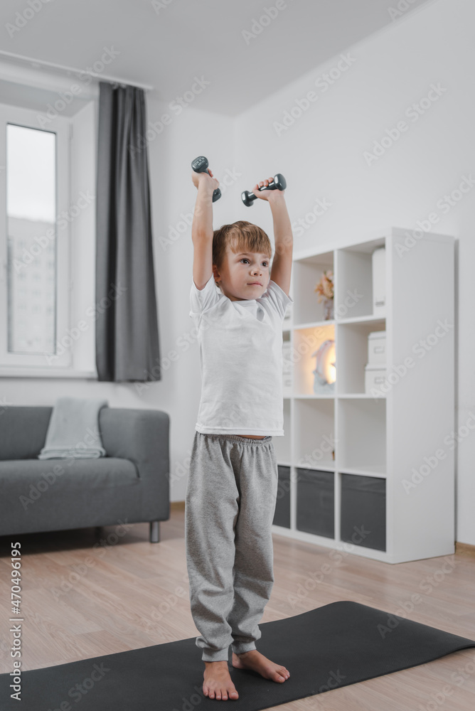 Sports hall at home. Boy child exercising with dumbbells in the living room. The child is dressed in a white t-shirt and gray pants. Sports hall at home. Boy child exercising with dumbbells in the living room. The child is dressed in a white t-shirt and gray pants.