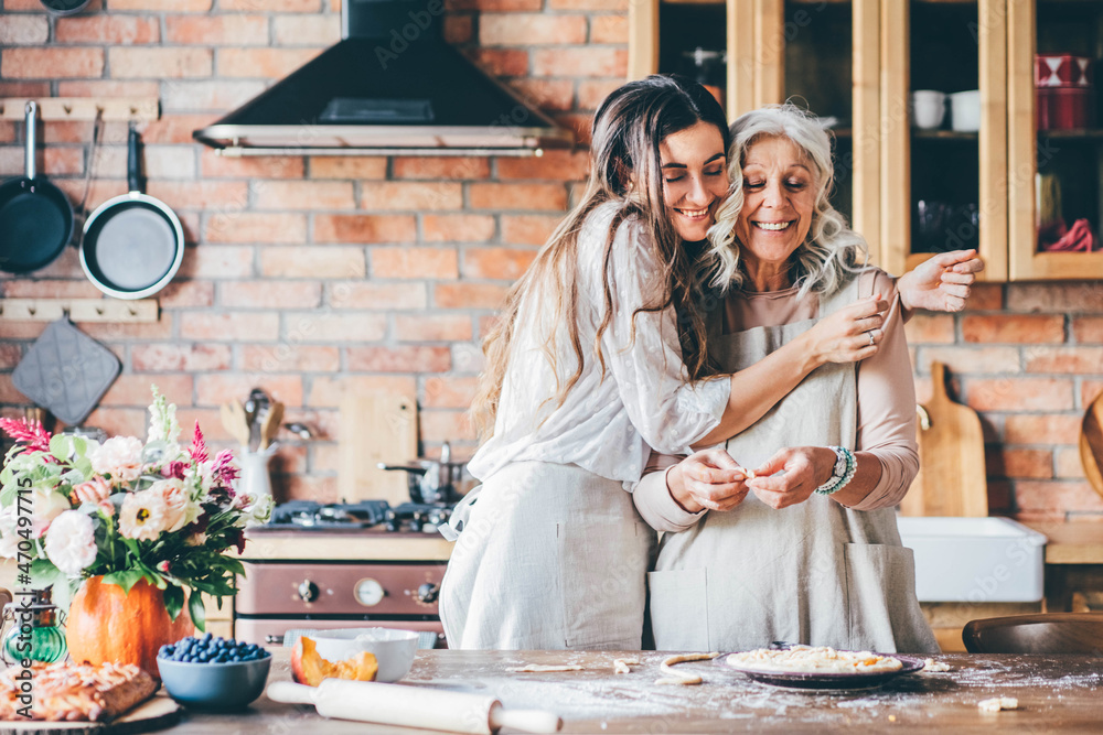 Young woman and mother in white aprons stand at large brown wooden kitchen table and prepare tasty dinner. Senior mother and happy adult daughter baking pumpkin pie together at home. - obrazy, fototapety, plakaty 