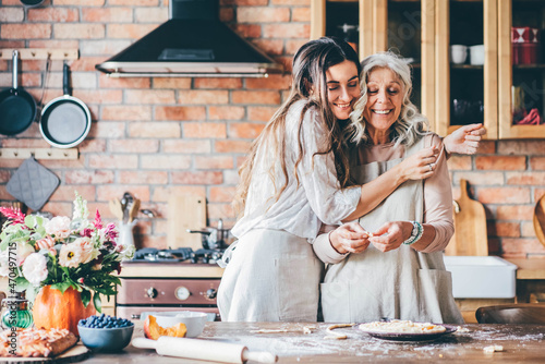 Fotografia, Obraz Young woman and mother in white aprons stand at large brown wooden kitchen table and prepare tasty dinner