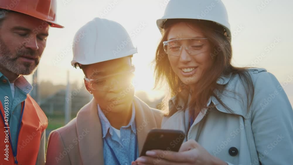 Trio of multi-ethnic professional architect with female engineers wearing helmets using mobile phone sharing screen communicating at power plant.
