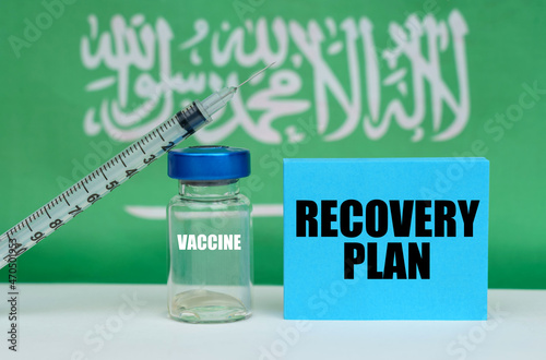 Vaccine, syringe and blue plate with the inscription - RECOVERY PLAN. In the background the flag of Saudi Arabia