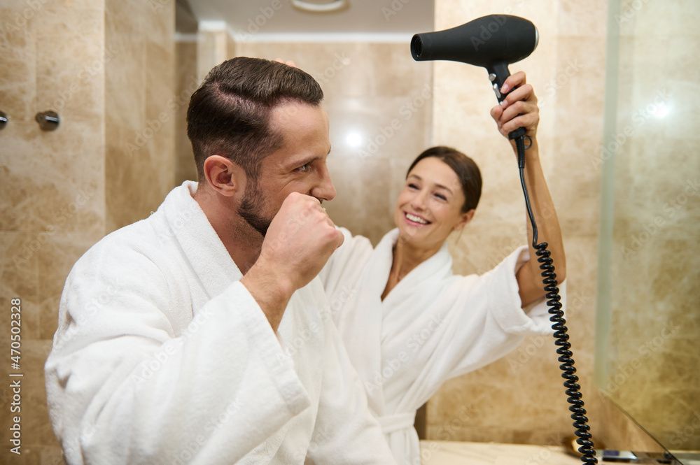 Handsome man in white bathrobe brushing teeth while his female partner smiles directing hair dryer on his hair. Happy middle aged couple in love enjoying morning routine together in bathroom Handsome man in white bathrobe brushing teeth while his female partner smiles directing hair dryer on his hair. Happy middle aged couple in love enjoying morning routine together in bathroom