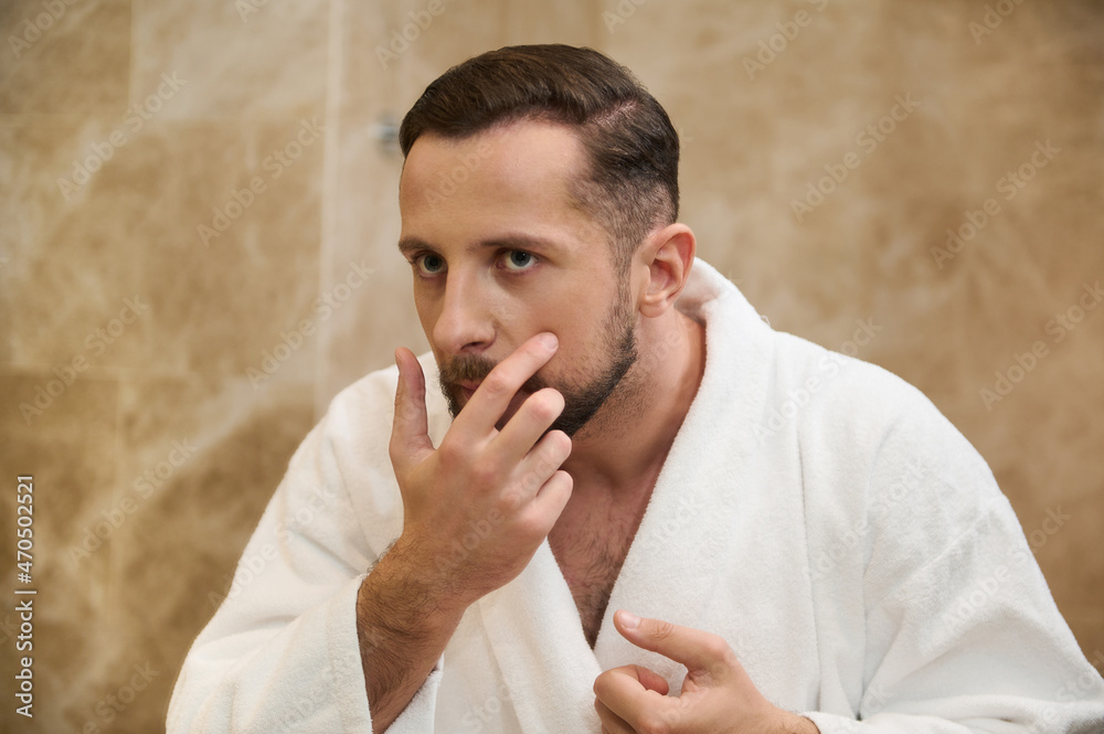 Headshot of a mirror reflection of a young man in white bathrobe worried about his appearance, looking at himself in the bathroom and examines his face for acne during morning hygiene routine Headshot of a mirror reflection of a young man in white bathrobe worried about his appearance, looking at himself in the bathroom and examines his face for acne during morning hygiene routine