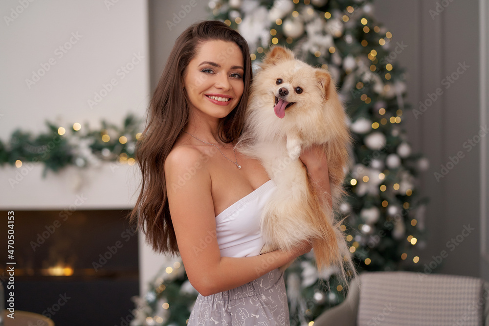 Young pretty woman holding small german spitz dog at her hands and standing against christmas tree with lights. Happy family Young pretty woman holding small german spitz dog at her hands and standing against christmas tree with lights. Happy family