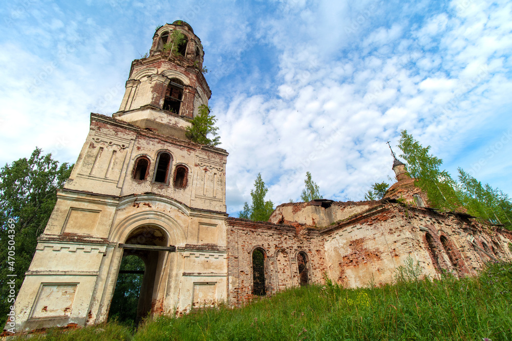 View of the front side of an old, destroyed and abandoned church in Russia. Walls with peeling paint and old red bricks. Trees on the roofs. Summer. Daylight. Sky with clouds.