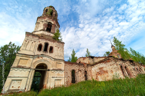 View of the front side of an old, destroyed and abandoned church in Russia. Walls with peeling paint and old red bricks. Trees on the roofs. Summer. Daylight. Sky with clouds.
