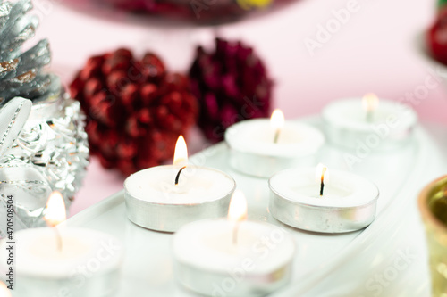 White Tealight Candles Illuminated In Row With Maroon Pine Cone Glitter Decorations On Light Pink Background. Theme For Merry Christmas And Happy New Year