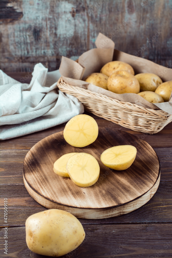 Fresh yellow potatoes cut into pieces on a board and a basket with tubers on a wooden table. Vegetarian food. Vertical view