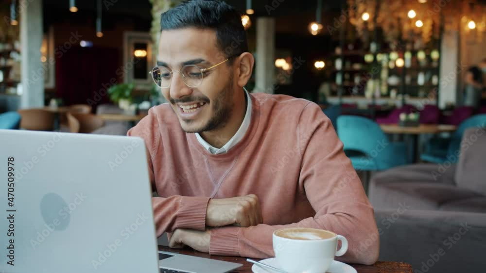 Portrait of happy young Middle Eastern man chatting on video call and waving hand using laptop in cafe. Communication and modern devices concept. Portrait of happy young Middle Eastern man chatting on video call and waving hand using laptop in cafe. Communication and modern devices concept.