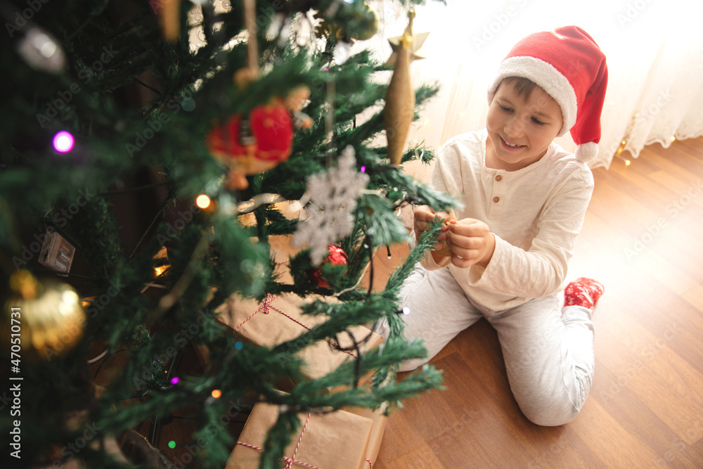 Smiling Caucasian boy in Santa's hat decorating Christmas tree sitting on the floor.