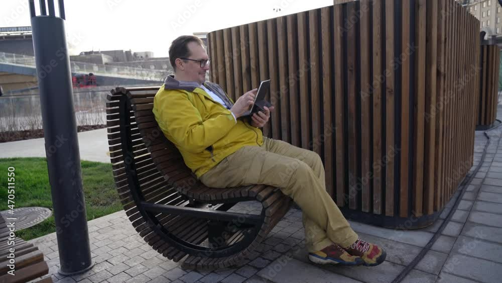 Millennial man in a yellow jacket is sitting on a bench in the city and reading work mail on a tablet against the background of wooden slats in the background Millennial man in a yellow jacket is sitting on a bench in the city and reading work mail on a tablet against the background of wooden slats in the background