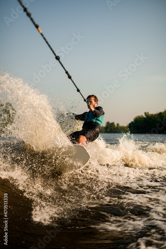 Athletic guy holds rope and speedly riding wakesurf board on splashing wave.