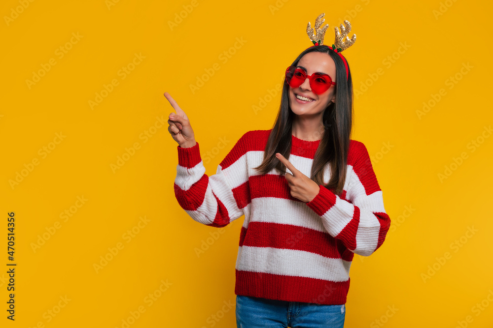 Close up portrait of an attractive excited smiling young girl in Christmas reindeer antlers and sweater while she pointing away on yellow background in studio Close up portrait of an attractive excited smiling young girl in Christmas reindeer antlers and sweater while she pointing away on yellow background in studio