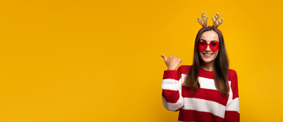 Banner wide photo of an attractive excited smiling young girl in Christmas reindeer antlers and sweater while she posing isolated on yellow background in studio