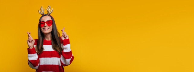 Banner photo of cute happy and charming young woman in a Christmas reindeer antlers with crossed fingers is praying for a miracle in New Year