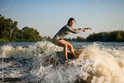 active flexible woman skillfully balancing on great splashing wave on wakesurf board. © fesenko