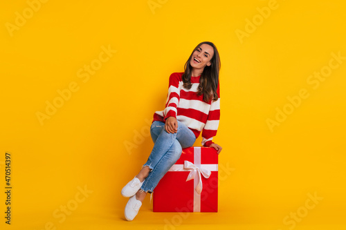 Full length photo of an excited beautiful stylish brunette woman while she is sitting and posing with a big red gift box
