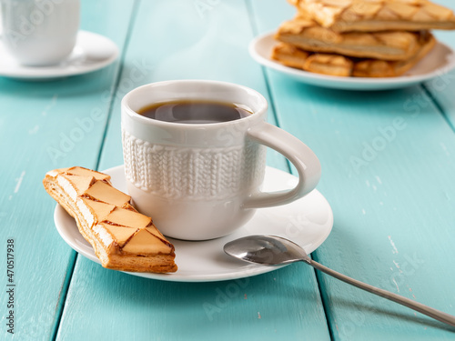 Cup of hot steaming coffe and Sfogliatine glassate biscuit on a white saucer over turquoise wooden table close-up. Sweet breakfast with italian crispy puff pastries and freshly brewed coffee. photo