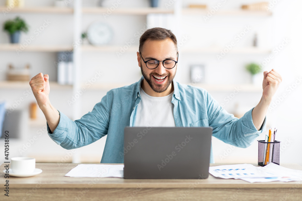 Young Caucasian guy gesturing YES in front of laptop, celebrating success at home office