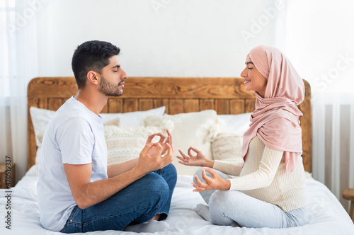 Pregnancy and yoga concept. Expectant muslim couple meditating together at home, sitting in lotus pose on bed