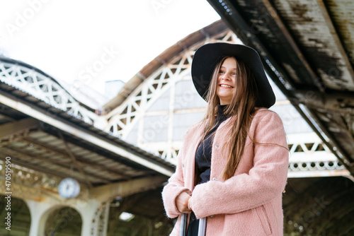 Beautiful young woman in black hat and pink coat with suitcase stands on platform of the station. Tourism and travel concept. Going on vacation or business trip
