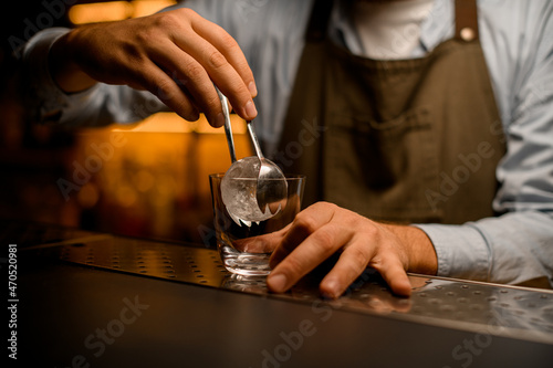 hand of bartender accurate holds round piece of ice by tongs and put it into glass