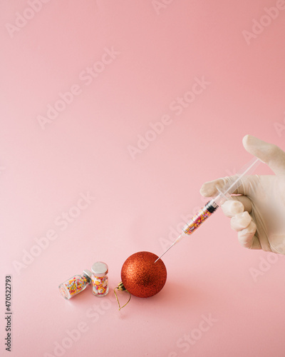 Covid inspired Christmas and new year design and concept. Vaccine in bottle needle red baubles with doctor hands in medical glows on colored background pink pastel.