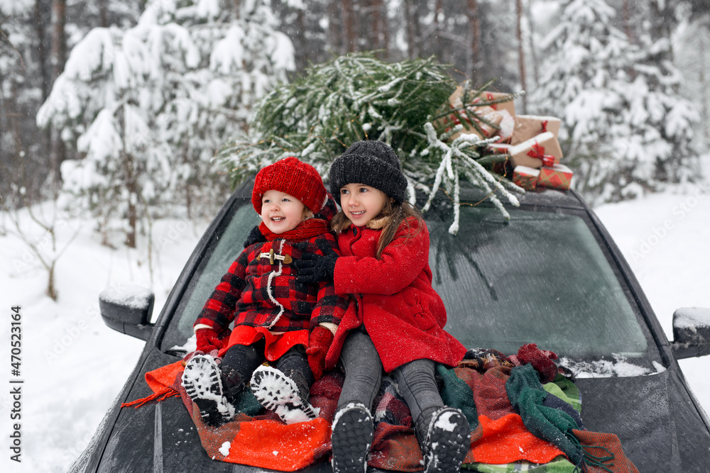 Cheerful children are sitting on the hood of a black car, on the roof of which there is a Christmas tree from the market and gifts with a red ribbon. Preparation for the celebration of New Year.