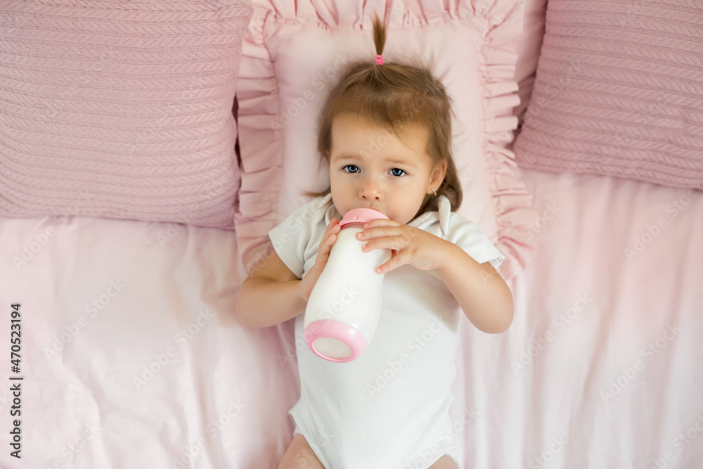Infant drinks a mixture of milk from a bottle lying on a bed with pink pillows. Healthy baby food for babies. Cute pretty small girl in a white bodysuit looks at the camera. Top view. Infant drinks a mixture of milk from a bottle lying on a bed with pink pillows. Healthy baby food for babies. Cute pretty small girl in a white bodysuit looks at the camera. Top view.