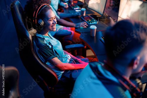 Cheerful African female cybersport gamer wearing wired headset sitting on gaming chair and watching game of teammate in cyber club