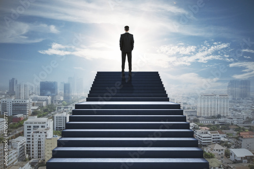 Back view of businessman standing on stairs on blurry city background. Success and growth concept. © Who is Danny