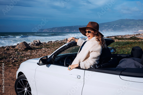 Outdoors lifestyle fashion portrait of pretty young woman driving cabriolet. Laughing and standing in car. Wearing stylish sheepskin coat, sunglasses, hat. Driving towards the ocean. Girl having fun