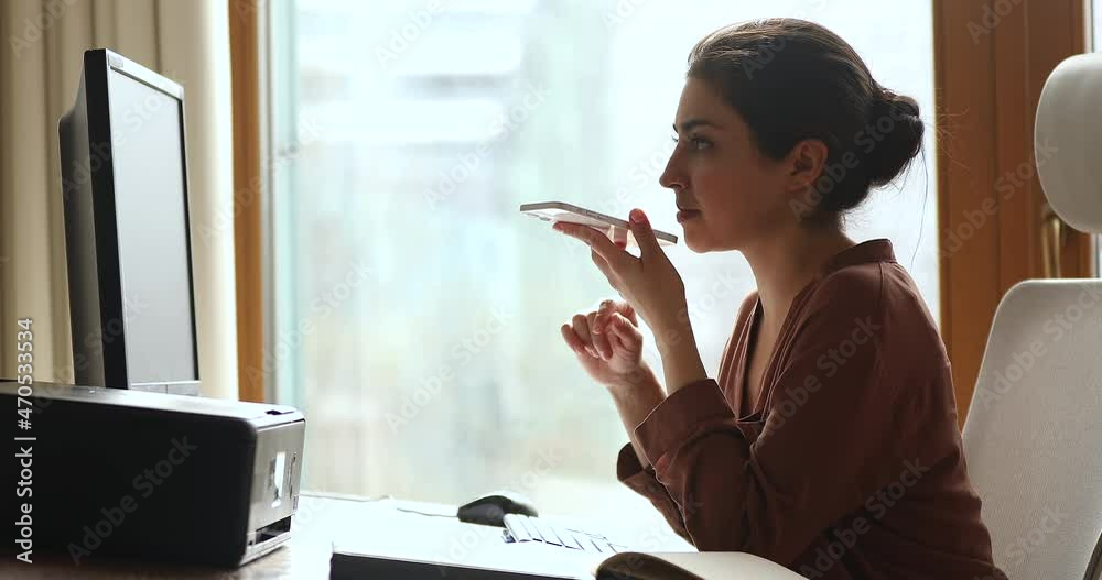 Confident young Indian businesswoman work by pc dictate vocal message using voice app on smartphone send voicemail to client business partner. Millennial lady office employee use ai assistant on phone
