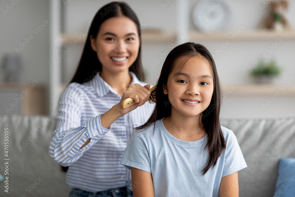 Loving asian mother combing her daughter hair with wooden brush while sitting together on sofa at home Loving asian mother combing her daughter hair with wooden brush while sitting together on sofa at home