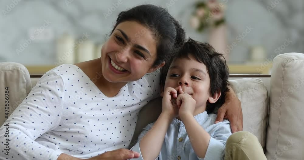 Cute little Indian boy sit on couch trust secrets to beloved mother nanny grownup elder sister. Young loving mom and little child cuddle on sofa whisper funny mysteries rumors on ears to each other Cute little Indian boy sit on couch trust secrets to beloved mother nanny grownup elder sister. Young loving mom and little child cuddle on sofa whisper funny mysteries rumors on ears to each other