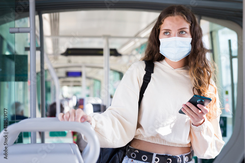 Young attractive woman in mask using mobile phone on way to work in modern tram