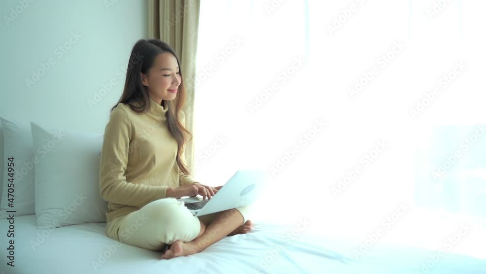 While sitting on a bed close to a large window, a young woman in a sweater and chinos sits cross-legged while working on her laptop. While sitting on a bed close to a large window, a young woman in a sweater and chinos sits cross-legged while working on her laptop.