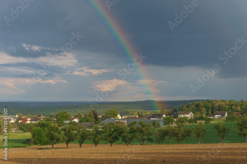 Detail of rainbow over a plowed field and trees with village in background, Germany