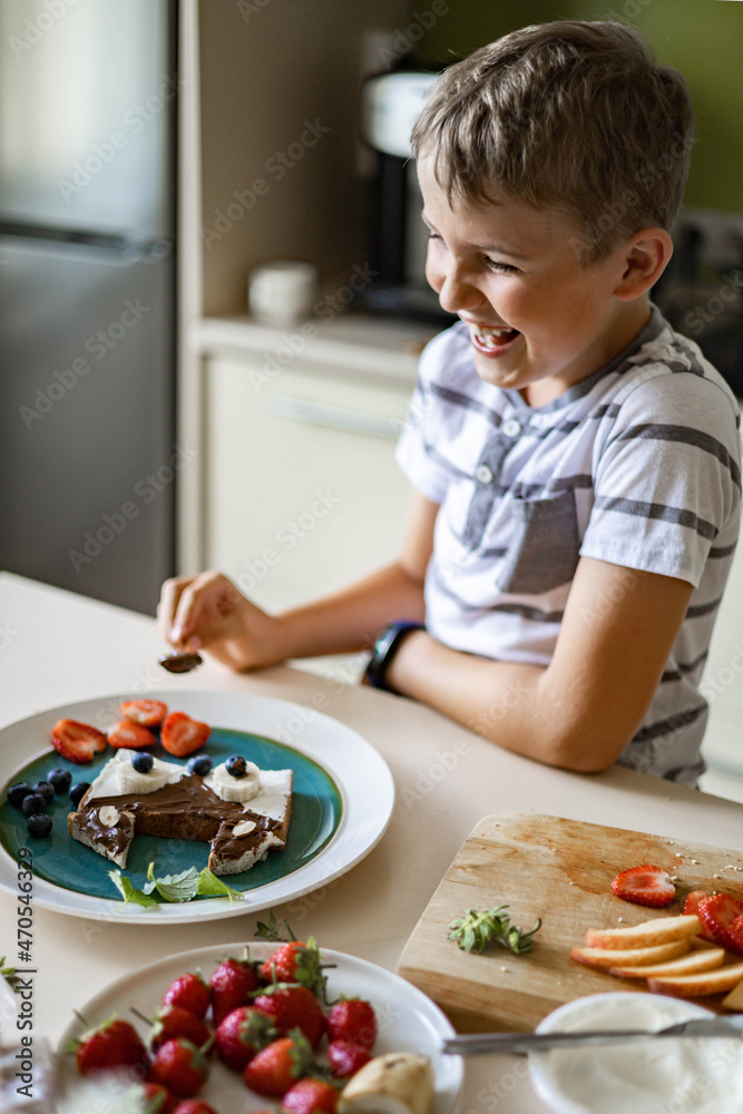 Laughing male kid cooking breakfast decorating fresh sandwich with berries and fruits at kitchen Laughing male kid cooking breakfast decorating fresh sandwich with berries and fruits at kitchen