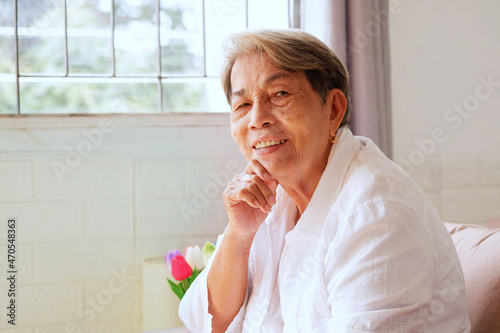 Elderly Asian woman with grey hair sits with her chin on the sofa with a happy smile on her face.