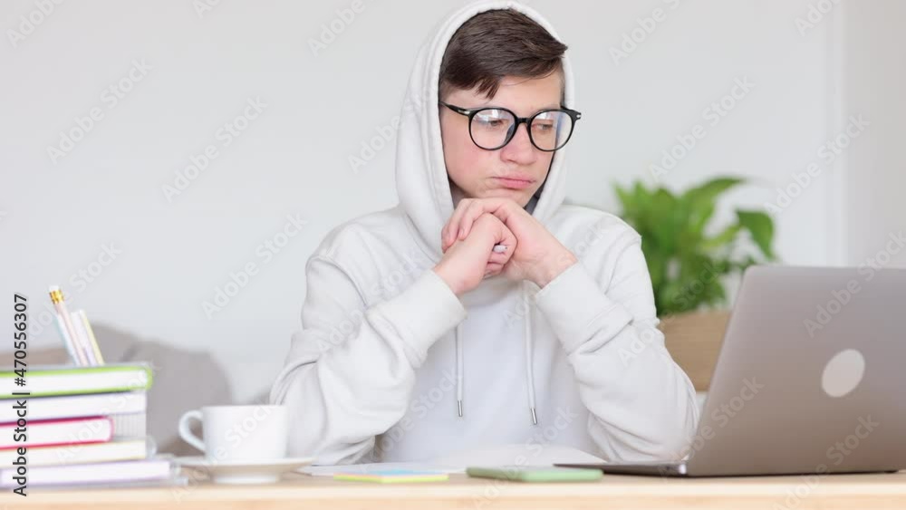 Online home school for teenage students. Calm teen schoolboy in white hoodie spbi watches bored video lesson via notebook computer at table in living room 4k video Online home school for teenage students. Calm teen schoolboy in white hoodie spbi watches bored video lesson via notebook computer at table in living room 4k video