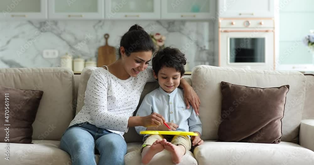 Loving single Indian mom engaged in drawing funny pictures on dry erase board sitting on couch at home hugging little boy preschooler. Friendly young mother teach small son to create simple sketches Loving single Indian mom engaged in drawing funny pictures on dry erase board sitting on couch at home hugging little boy preschooler. Friendly young mother teach small son to create simple sketches